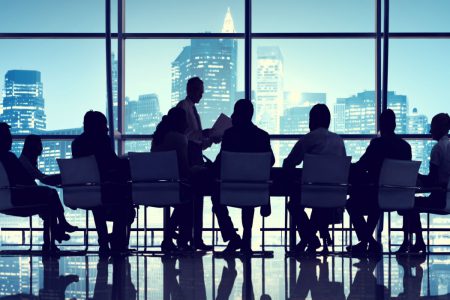 An office meeting at a long table in front of a dark city landscape.