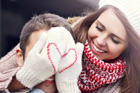 A woman hiding a man's face with wollen gloves with a heart pattern on the front on valentines day