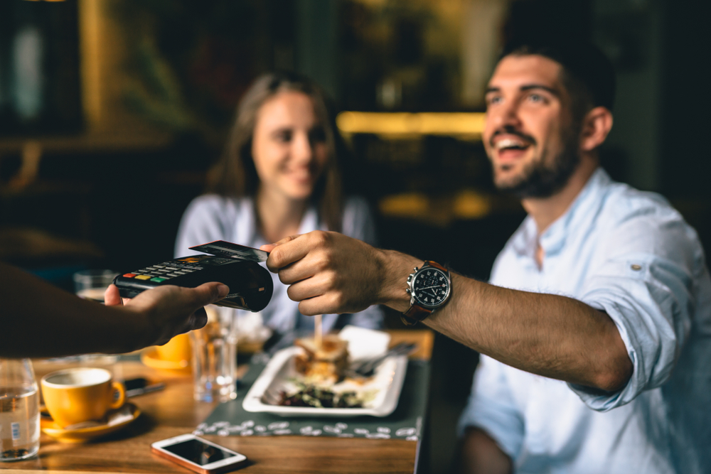 man paying bill at a restaurant, tapping bank card onto contactless payment machine. Burger on the table in front of him and a woman enjoying her meal.