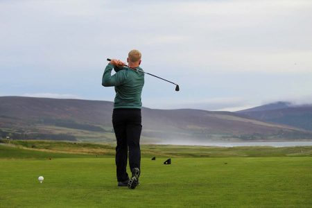A golfer in a green jumper follows through on a tee shot, with heather-covered hills and the sea behind him.