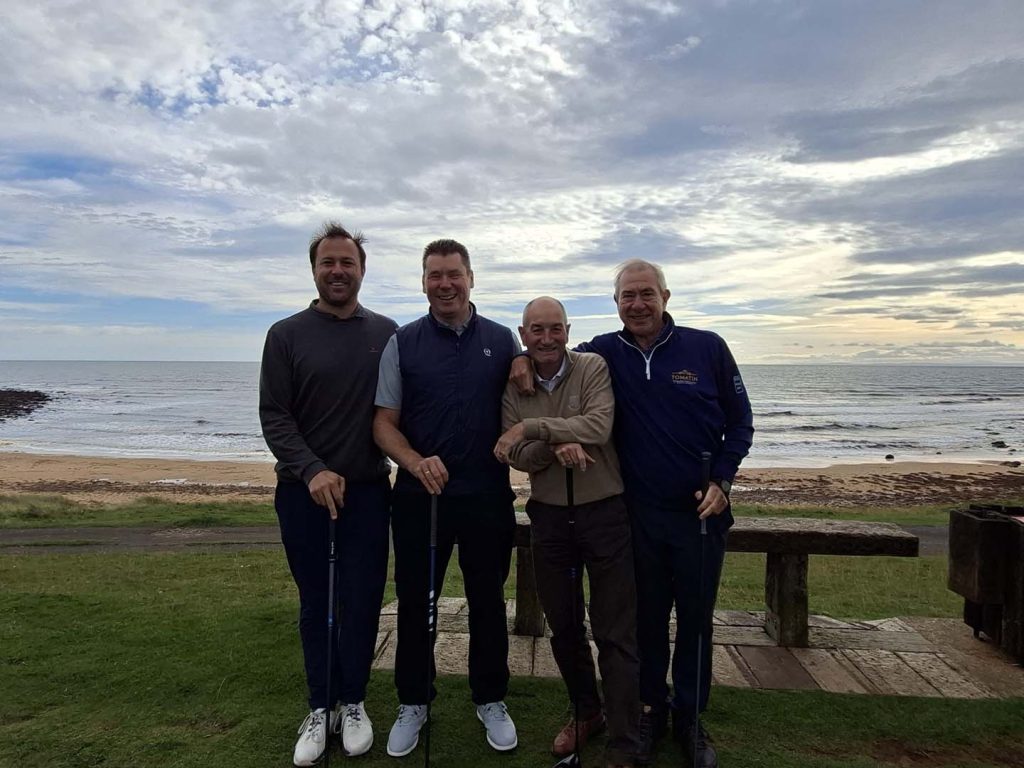 Four golfers stand smiling with their clubs on a grassy tee, with a sandy beach and the sea behind them.