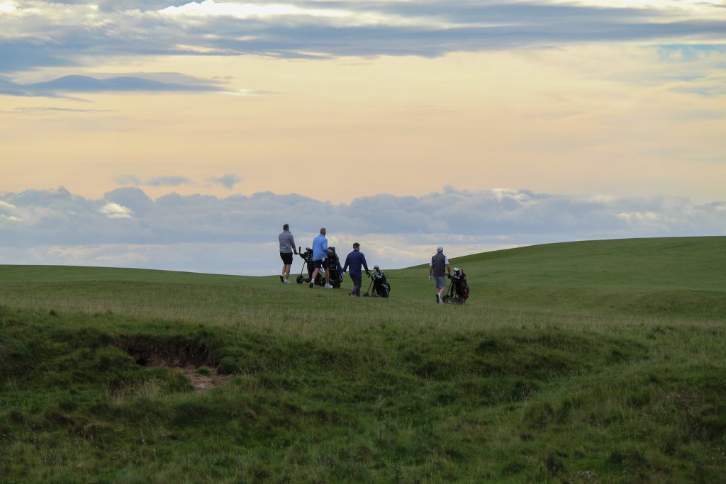 Four golfers pull their trolleys across a rolling links fairway under a pale golden evening sky.