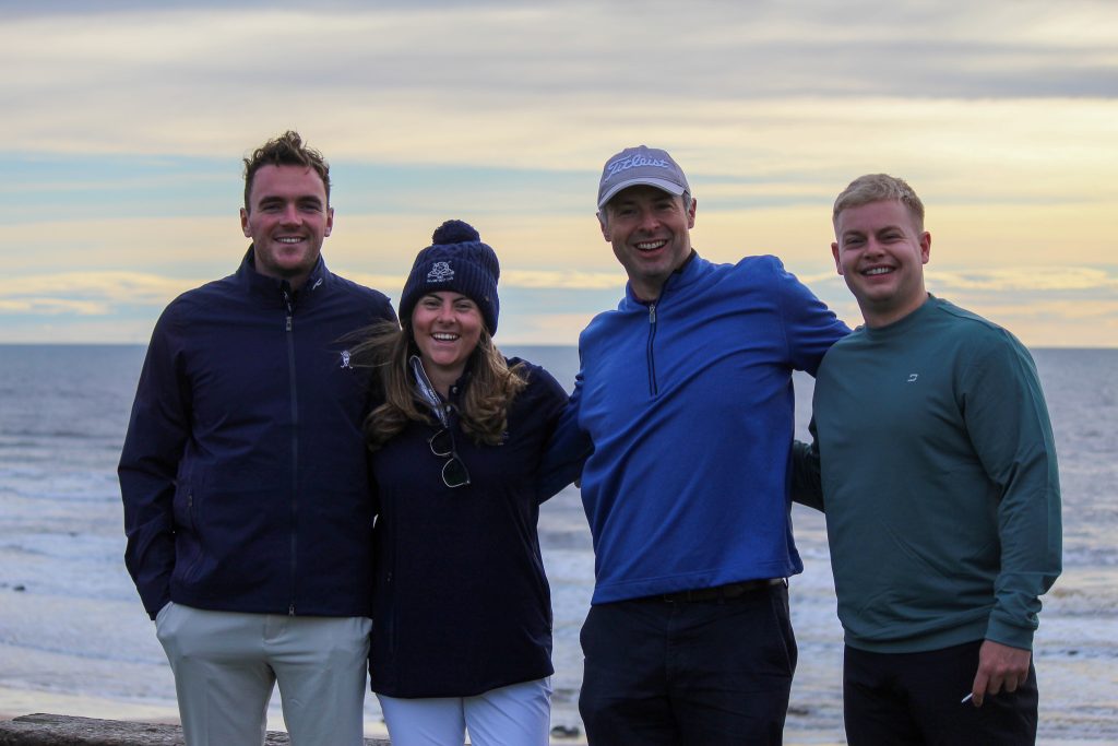 Four smiling golfers, one in a bobble hat, pose arm in arm by the sea at sunset.