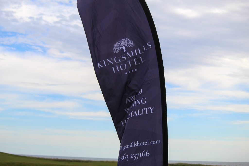 A purple Kingsmills Hotel feather flag reading “Award Winning Hospitality” flutters against a blue sky over the links.